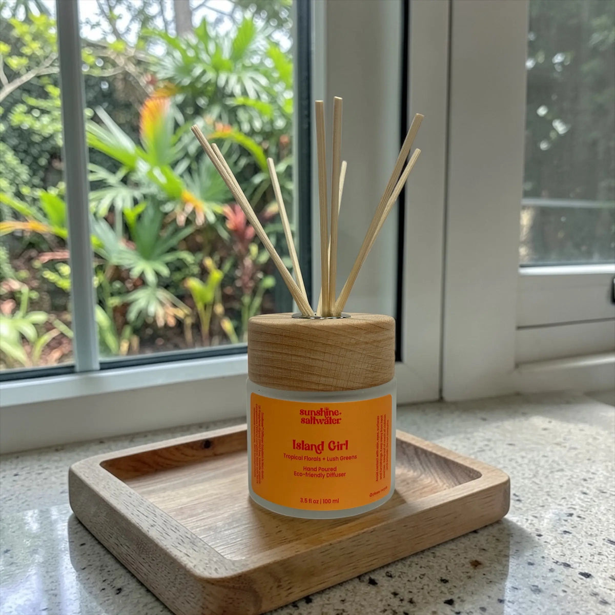 Reed diffuser with wooden lid on a wooden coaster by a window with tropical greenery and plants outside