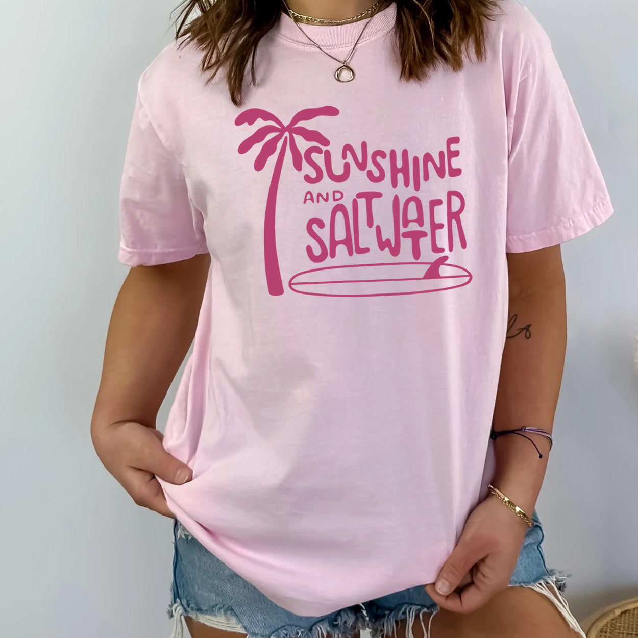 bright pink t-shirt with dark pink sunshine and saltwater graphic next to palm tree