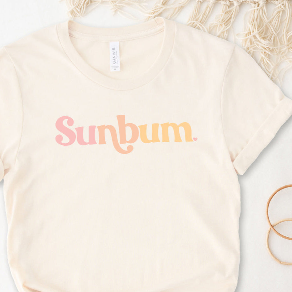 A beige t-shirt with the text &#39;Sunbum&#39; printed in a pinkish hue, displayed flat with a textured object to the left.