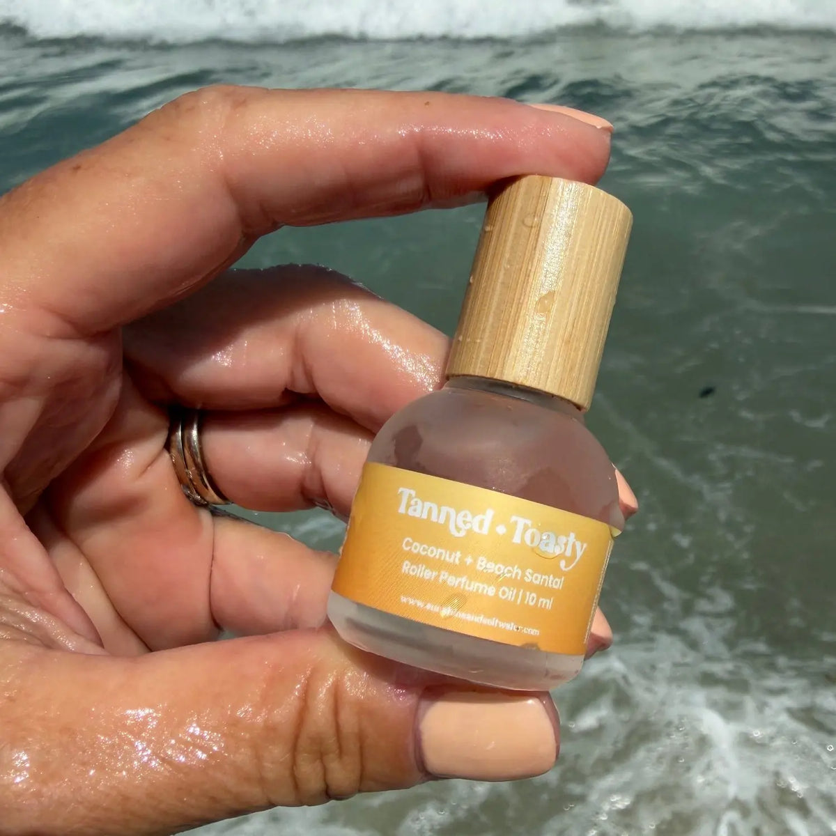 A hand holding a small bottle of Tanried + Truly Coconut + Santal rollerball against a beach background with ocean waves.