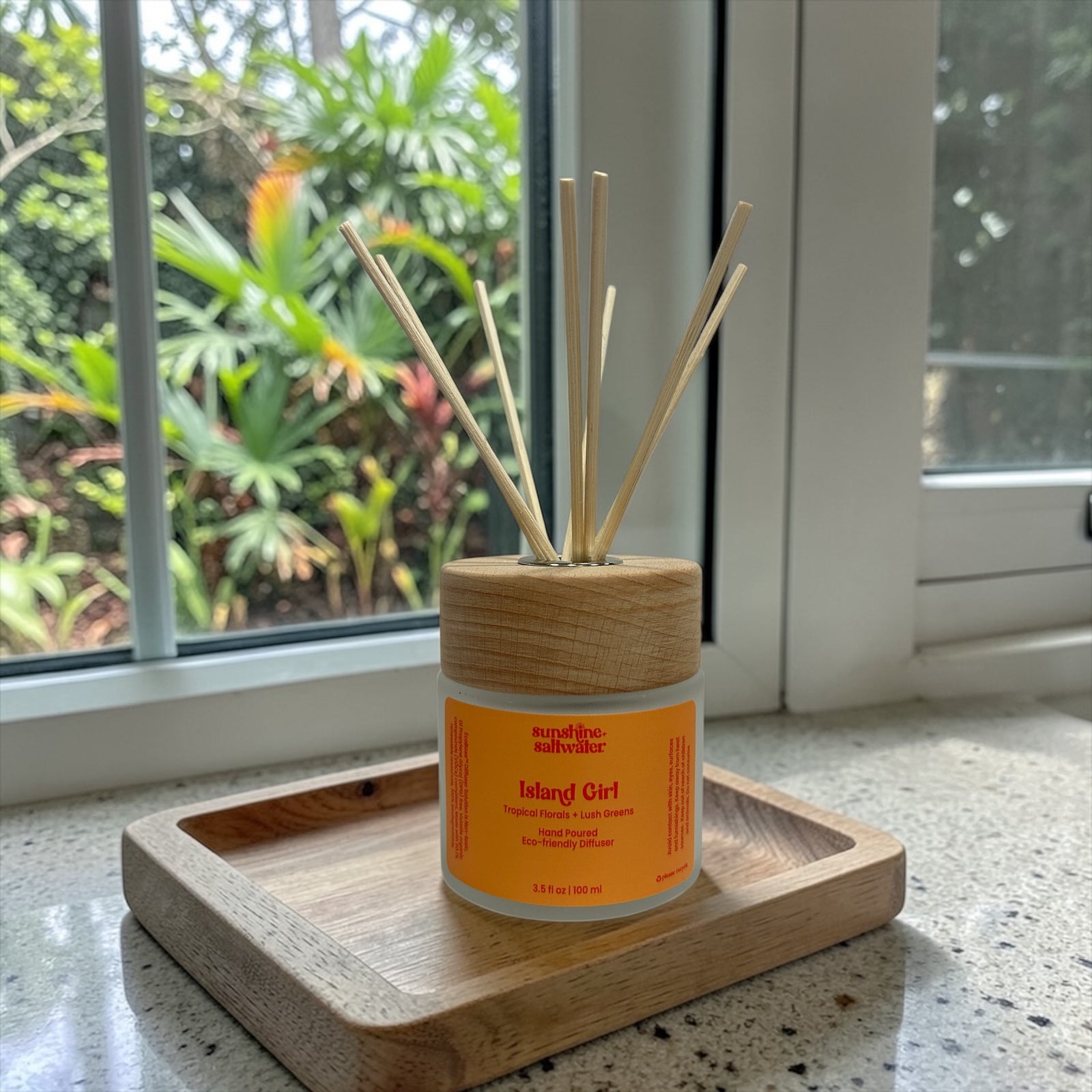 A tropical floral and lush greens diffuser with sticks on a wooden tray in front of a window with tropical greenery outside.