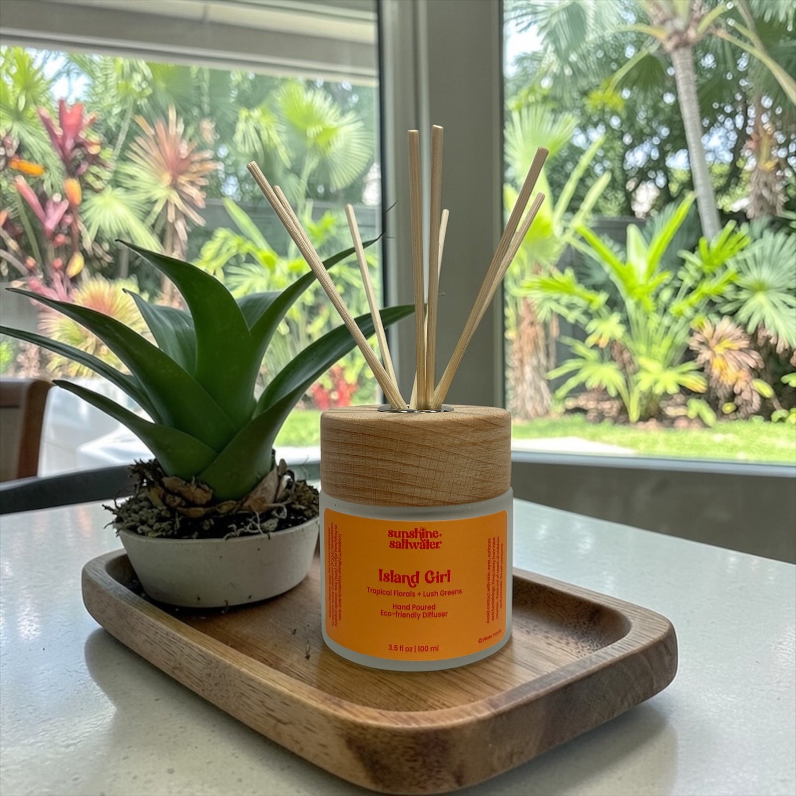 A tropical floral and lush greens diffuser with sticks on a wooden tray in front of a window with tropical greenery outside.