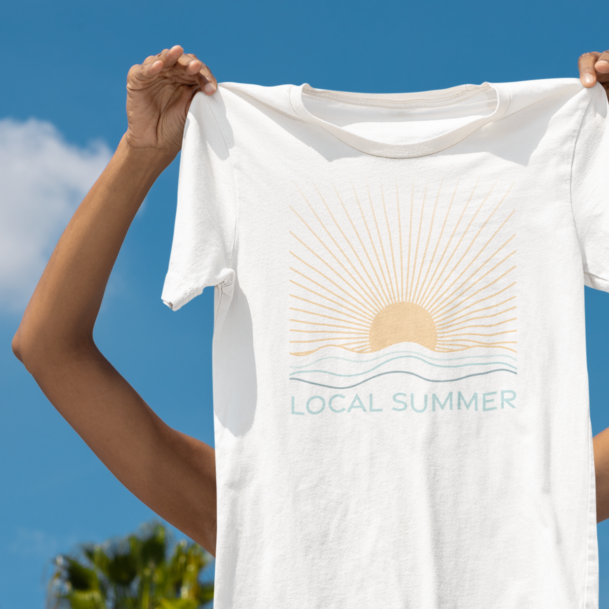 Person holding a white t-shirt with &#39;Local Summer&#39; design against a blue sky.